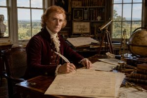Thomas Jefferson at his Monticello study writing desk with quill pen working on Declaration of Independence manuscript, surrounded by telescope, globe, architectural drawings, and books, wearing burgundy 18th century coat with white cravat, Virginia countryside visible through large windows